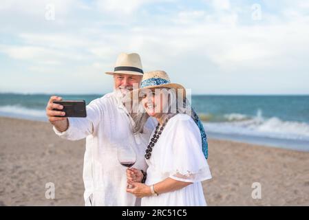 Positives älteres Paar in weißen Strandkleidung, das Selfie mit dem Smartphone machte, während es am Sandstrand in der Nähe des Wellenmeers stand Stockfoto