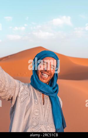 Lächelnder junger Berber in traditioneller Kleidung und blauem Turban, der Selfie mit dem Smartphone in der Merzouga-Wüste Marokko macht Stockfoto