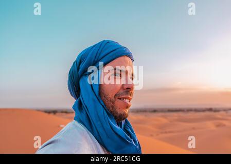 Seitenansicht eines glücklichen Berbers mit blauem traditionellem Turban, der wegblickt, während er in der Wüste von Merzouga vor blauem Himmel steht Stockfoto