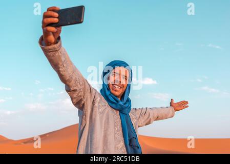 Fröhlicher junger Berber in traditioneller Kleidung und blauem Turban, der Selfie mit dem Smartphone in der Merzouga-Wüste Marokko macht Stockfoto