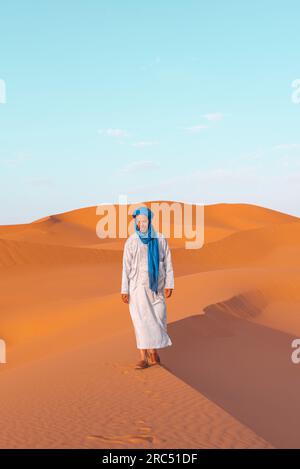 Ein ganzer Körper junger Berber in traditioneller Tuareg-Kleidung, der auf einer Sanddüne spaziert, während er in die Merzouga-Wüste Marokko blickt Stockfoto