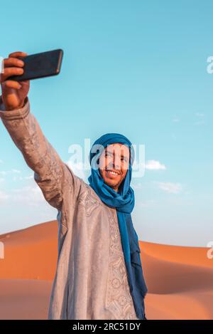 Lächelnder junger Berber in traditioneller Kleidung und blauem Turban, der Selfie mit dem Smartphone in der Merzouga-Wüste Marokko macht Stockfoto