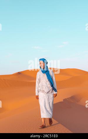 Ein ganzer Körper junger Berber in traditioneller Tuareg-Kleidung, der auf einer Sanddüne spaziert, während er in die Merzouga-Wüste Marokko blickt Stockfoto