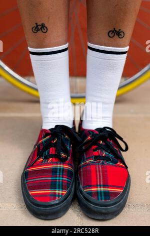 Anonyme Frau in weißen schwarzen Socken und Turnschuhen mit temporären Tattoos kleiner Fahrräder auf Beinen unterhalb des Knies gegen gelbes Rad Stockfoto