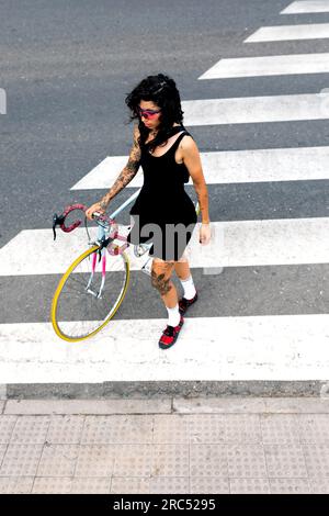 Junge hispanische Frau mit roter Sonnenbrille Sneaker Socken schauen weg, halten den Griff des Fahrrads und laufen tagsüber auf der Straße über Zebrastreifen Stockfoto