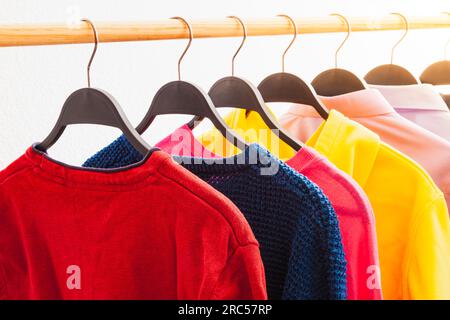 Mehrere langärmelige Kleidungsstücke in verschiedenen Farben hing an Kleiderbügeln auf einer runden Holzstange. Im Hintergrund befindet sich eine weiße Wand. Stockfoto