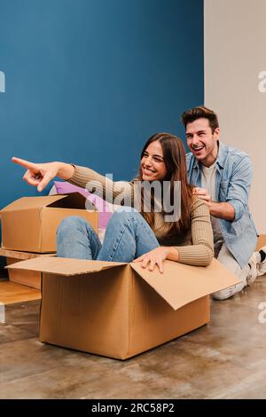 Vertikales Porträt eines Paares, das beim Auspacken in seiner neuen Wohnung spielt. Glückliche Frau sitzt in einer Pappschachtel und amüsiert sich mit ihrem lächelnden Freund, während sie zusammen umzieht. Hochwertiges Foto Stockfoto