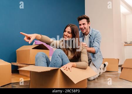 Fröhliches Paar, das beim Auspacken in seiner neuen Wohnung spielt. Aufgeregte und glückliche Frau, die in einer Pappschachtel sitzt und mit ihrem lächelnden Freund Spaß hat, während sie zusammen umzieht. Hochwertiges Foto Stockfoto