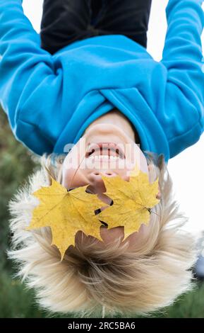 Porträt eines süßen, lustigen, blonden Jungen, der kopfüber hängt, zwei gelbe Ahornblätter statt Augen. Herbststimmung. Glückliche Kindheit. Freude, Verwöhnung, Spaß Stockfoto