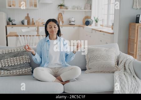 Eine heitere Frau übt Yoga, meditiert auf der Couch in Lotusposition. Atemübungen für mentale Balance, Angstlinderung. Gesunder Lebensstil zu Hause. Stockfoto