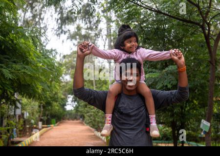 Ein liebevoller Vater trägt seine Tochter auf seinen Schultern, während sie durch den Park laufen. Der Vater lächelt und die Tochter lacht, und das sind sie Stockfoto