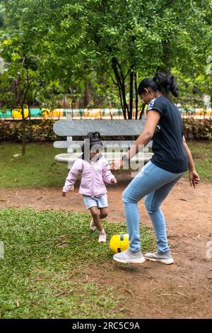 Ein glückliches 5-jähriges Mädchen und ihre Mutter spielen Fußball im Park. Das Mädchen trägt ein pinkfarbenes Fußballtrikot und Shorts, und ihre Mutter trägt sie Stockfoto
