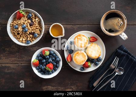 Frühstückstisch mit Morgenkaffee, Granola Schüssel und Hüttenkäse-Frittern serviert mit frischen Sommerbeeren und Honig. Draufsicht, Tischrücken aus Holz Stockfoto