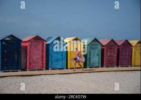 Alicante, Spanien. 12. Juli 2023. An einem heißen Sommertag am San Juan Beach führt ein Mann an einigen bunten Strandhütten vorbei, die einen aufblasbaren Schwimmer tragen. Die Staatliche Meteorologische Agentur (AEMET) hat rote Warnungen für hohe Temperaturen aktiviert, die durch eine Hitzewelle verursacht werden, die in einigen Gebieten Temperaturen über 40 Grad Celsius ansteigen wird. Kredit: Marcos del Mazo/Alamy Live News Stockfoto