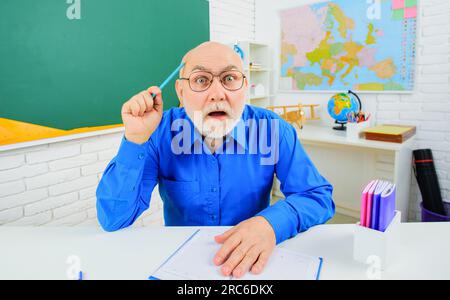 Überraschter bärtiger Lehrer am Schreibtisch im Klassenzimmer. Wissenstag. Bärtiger Lehrer, der sich auf die Prüfung im College vorbereitet. Lehrerprofessor im Unterricht Stockfoto