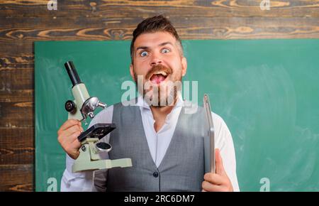 Lustiger College-Lehrer mit Computer und Mikroskop. Aufgeregter Lehrer mit Notebook und Mikroskop im Klassenzimmer. Wissenschaftler mit Laptop und Mikroskop Stockfoto