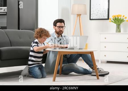 Mann, der zu Hause arbeitet. Vater benutzte Laptop, während sein Sohn am Schreibtisch zeichnete Stockfoto