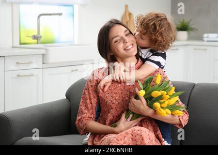 Der kleine Sohn küsst und gratuliert seiner mutter mit Muttertag zu Hause. Eine Frau mit einem Strauß gelber Tulpen Stockfoto