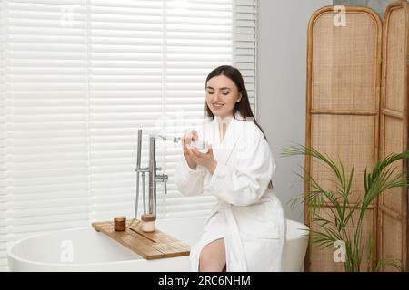 Eine schöne Frau im weißen Bademantel, die im Badezimmer Creme aufträgt Stockfoto