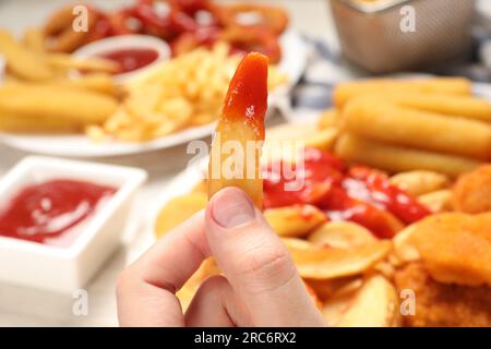 Eine Frau, die leckeren gebackenen Kartoffelkeil mit Ketchup in der Nähe verschiedener Snacks am Tisch hält, Nahaufnahme Stockfoto