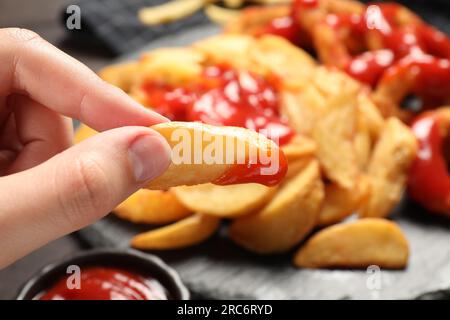 Eine Frau, die leckeren gebackenen Kartoffelkeil mit Ketchup in der Nähe verschiedener Snacks am Tisch hält, Nahaufnahme Stockfoto