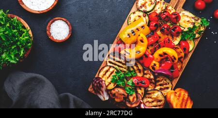Gegrilltes Gemüse auf rustikalem Holzschneidbrett: Bunte Paprika, Zucchini, Aubergine, Pilze, Tomaten und rote Zwiebeln mit Joghurt-Dressing, Bla Stockfoto