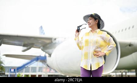 Am Flughafen steht ein Porträt einer attraktiven asiatischen Seniorin in einem Sommerkostüm mit Strandhut und Sonnenbrille. Sommerferienkonzept Stockfoto