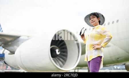 Eine attraktive ältere Asiatin in einem Sommeranzug mit Strandhut und Sonnenbrille steht am Flughafen. Sommerferienkonzept Stockfoto