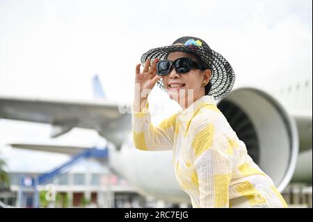 Am Flughafen steht ein Porträt einer attraktiven asiatischen Seniorin in einem Sommerkostüm mit Strandhut und Sonnenbrille. Sommerferienkonzept Stockfoto