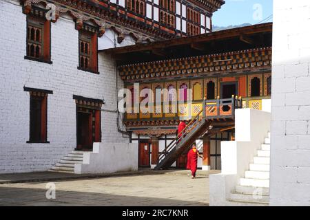Zwei buddhistische Mönche mit roten Rändern gehen in der Nähe eines traditionellen, bunt bemalten Gebäudes auf dem Gelände von Tashichho Dzong in Thimphu, Bhutan. Stockfoto