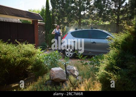 Ein junger Mann in einem Arbeitsanzug wäscht sein Auto mit einem Wasser-Hochdruckreiniger im Garten Stockfoto