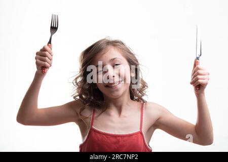 Ein kleines Mädchen in einem roten Tanktop, das mit Gabel und Messer gespannt auf das Essen wartet, wartet freudig auf sie Stockfoto