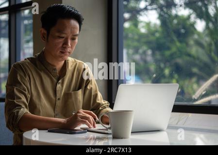 Ein asiatischer Mann, der mit seinem Laptop und Smartphone auf dem Schreibtisch arbeitet, konzentrierte sich auf seine Arbeit, mit einem ernsten Gesichtsausdruck. Stockfoto