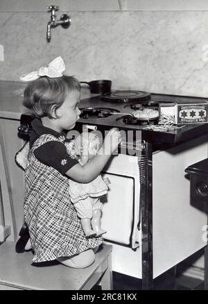 Vereinigte Staaten: ca. 1932 Ein kleines Mädchen hält ihre Puppe und versucht, einen Gasherd mit Streichhölzern anzuzünden, um einen Keks zu kochen. Stockfoto