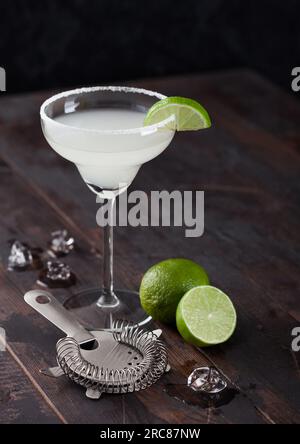 Klassisches Glas Margarita-Cocktail mit frischen Limetten und Sieb mit Eiswürfeln auf Holztisch. Stockfoto