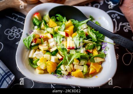 Obst- und Gemüsesalat in Schüssel mit Löffel auf schwarzem Tisch, Nahaufnahme. Orangenscheiben, Apfel, Fenchel und Gurken mit Rucola-Blättern. Stockfoto