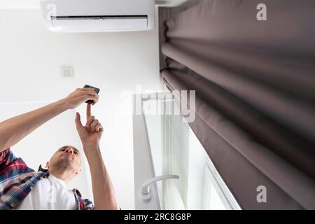 Langer junger Mann mit Brille, der die Fernbedienung der Klimaanlage benutzt, entspannt, auf der Couch sitzt, umschaltet, bequem einstellen Stockfoto