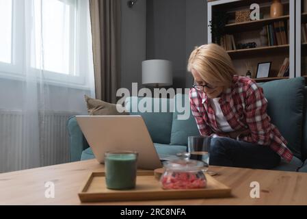 Müde, gestresst, verärgert, Frau mittleren Alters, krank, Kopfschmerzen nach der Computerarbeit, sitzen zu Hause auf dem Sofa, traurige, ermüdete alte Seniorenmutter Stockfoto