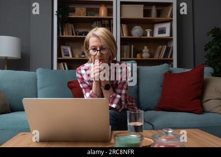Müde, gestresst, verärgert, Frau mittleren Alters, krank, Kopfschmerzen nach der Computerarbeit, sitzen zu Hause auf dem Sofa, traurige, ermüdete alte Seniorenmutter Stockfoto