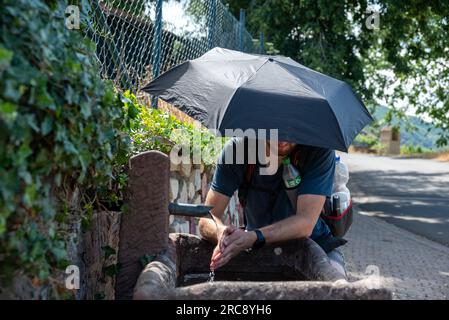 Neumagen Dhron, Deutschland. 13. Juli 2023. Ein Wanderer mit Sonnencreme erfrischt sich an einem Brunnen entlang einer Farm. Im Kampf gegen die Gesundheitsrisiken durch Hitze hat die Landesregierung Rheinland-Pfalz eine Website mit Tipps und Informationen zum Verhalten gestartet. Trinken Sie viel Flüssigkeit, meiden Sie die Sonne, tragen Sie helle Kleidung und schützen Sie Ihren Kopf. Kredit: Harald Tittel/dpa/Alamy Live News Stockfoto