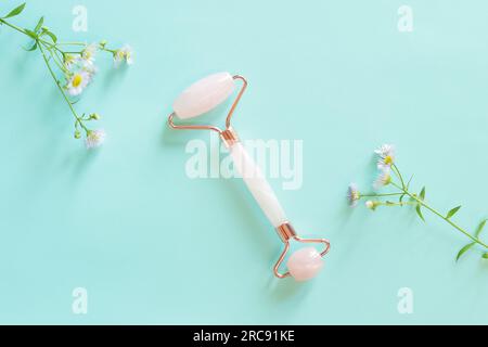 Gesichtsmassagewalze aus Rosenquarz auf blauem Hintergrund mit Kamillenblüten. Gesichtsmassagegerät Anti-Falten Anti-Aging-Hautpflegewerkzeug. Flachla Stockfoto