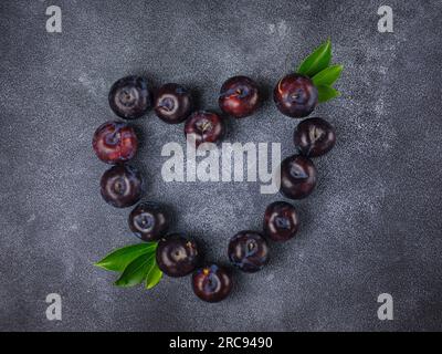 Süße Pflaumen auf dunklem Hintergrund. Frische blaue Pflaumen mit Blättern. Kulinarische Frucht im Hintergrund. Sommerpostkarte. Die Pflaumen werden im Garten geerntet und liegen in Herzform, Draufsicht Stockfoto