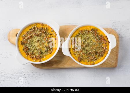 Hausgemachter Kürbisauflauf oder Gratin mit Käse und Kürbiskernen in zwei Keramikdosen auf hellgrauem Hintergrund, flach gelegt Stockfoto