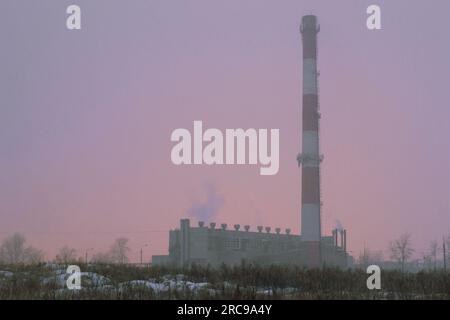 Der Kamin und das Gebäude des Kesselhauses im Nebel während eines Sturms vor dem Hintergrund des Lichts der Gewächshäuser Stockfoto