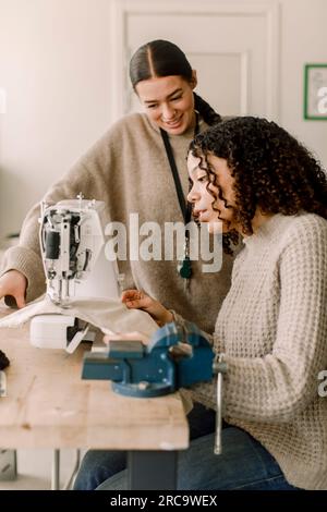 Lächelnde Lehrerin, die eine Schülerin ansieht, die eine Nähmaschine benutzt, in der Kunststunde der Highschool Stockfoto