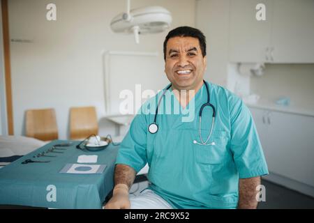 Porträt eines lächelnden männlichen Arztes, der Peelings trägt und im Untersuchungsraum sitzt Stockfoto