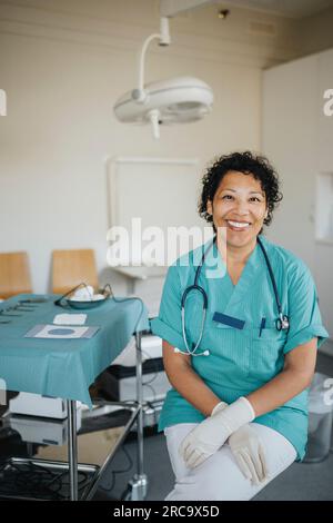 Porträt einer glücklichen Ärztin, die medizinische Peelings trägt und im medizinischen Untersuchungsraum sitzt Stockfoto