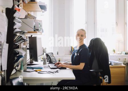Porträt einer lächelnden Gesundheitsarbeiterin, die am Schreibtisch in der Klinik sitzt Stockfoto