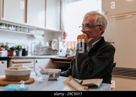 Rücksichtsvoller Senior-Mann mit Hand am Kinn, der zu Hause in der Küche sitzt Stockfoto