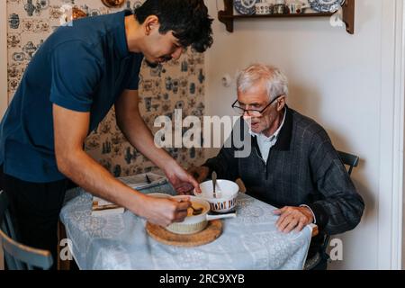 Männlicher Betreuer, der dem Seniorenmann, der zu Hause am Esstisch sitzt, Essen serviert Stockfoto